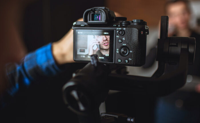 Picture of cameraman's hand adjust lens. Camera filming or taking picture of young man talking on phone. Recording video or taking photo. Blurred background.